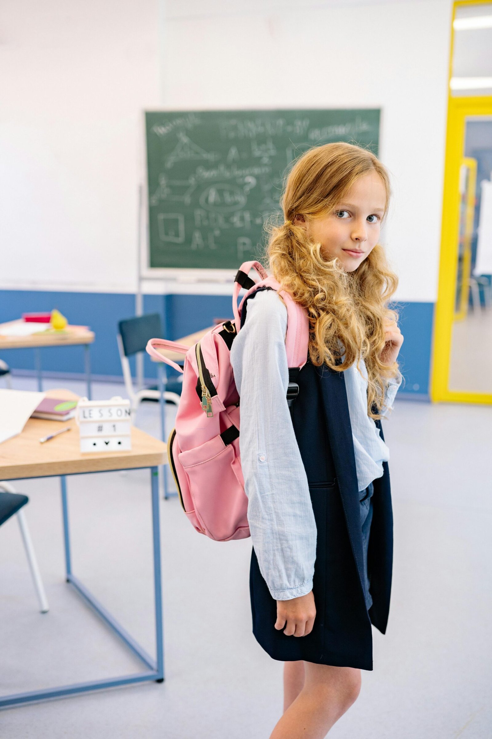 A young girl with a backpack stands confidently in a bright classroom setting.